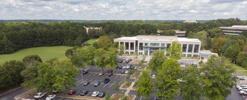 Aerial view of Johns Creek City Hall. A large building and parking lot surrounded by green lawn and lots of trees.