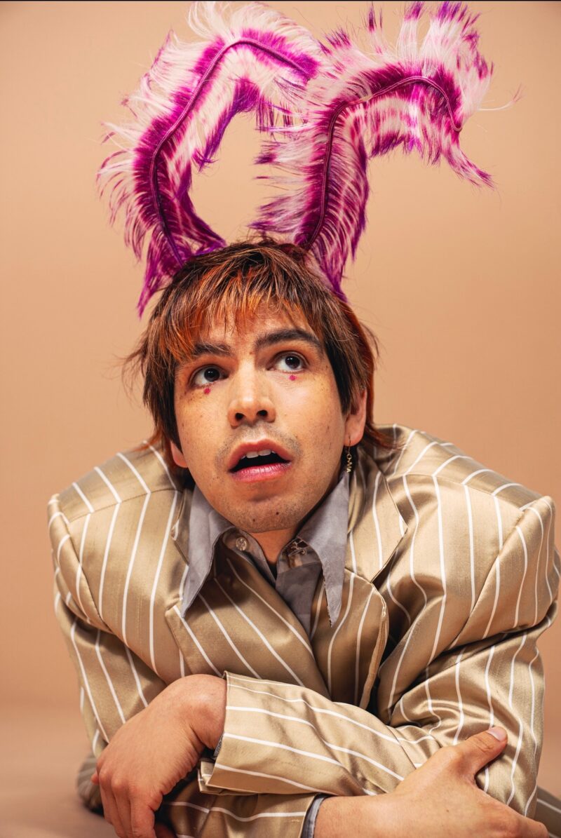 Julio Torres in festive headdress, a striped beige and white suit and makeup.