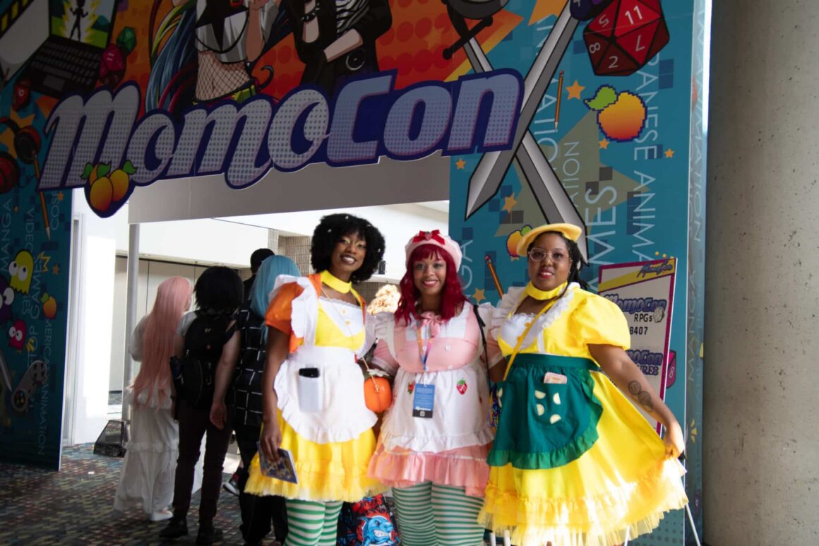 Three young Black women are cosplaying in cute anime costumes standing in front of the entrance to MomoCon in 2025.