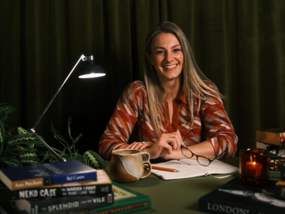 A young woman with long blonde hair is sitting at a desk in a low-lit room. There is a desk lamp, papers, pens, books and other items on the desk in front of her.