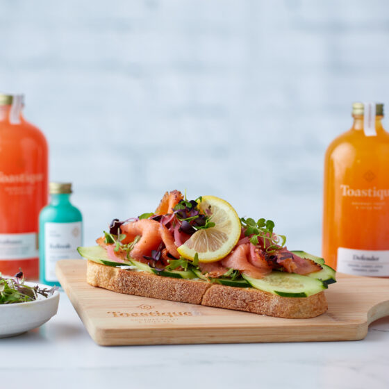 Smoked salmon toast on a wooden carving board with bottles of juice, wellness shots and other items. They are on a white table against a white background.