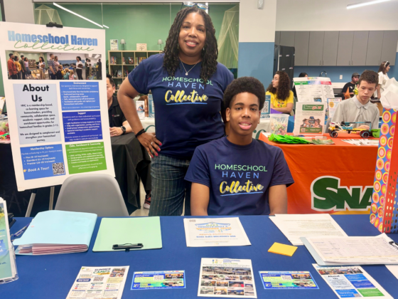 A Black woman and her teenage son are behind a table with a blue tablecloth and papers and brochures, welcoming families to the Homeschool Haven Collective that they founded.