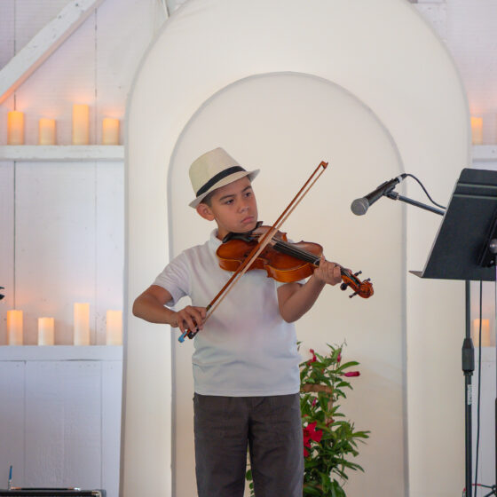 A young boy in a white shirt, dark pants and white trilby-style hat is standing behind a mic, playing violin. The background is a white wall with arch detailing and rows of white, lit candles.