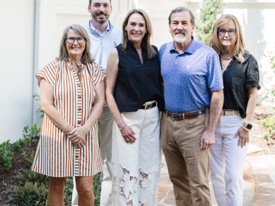 Five people who work together at a real estate agency are dressed nicely, standing outside next to a white house with white fence.