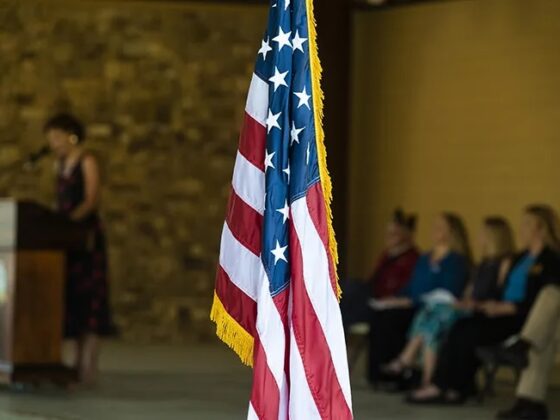 An American flag in the foreground with people sitting in chairs on a stage and one person at a podium in the background.