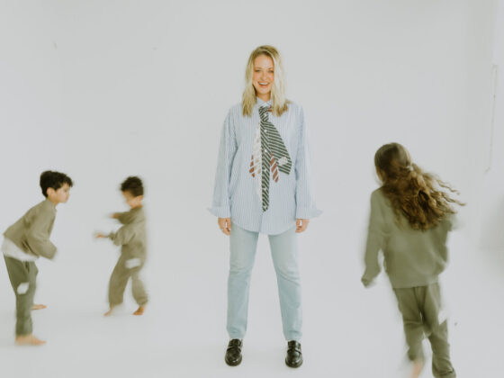 A blonde haired woman standing in the middle of three young children running around her against a white floor and background.