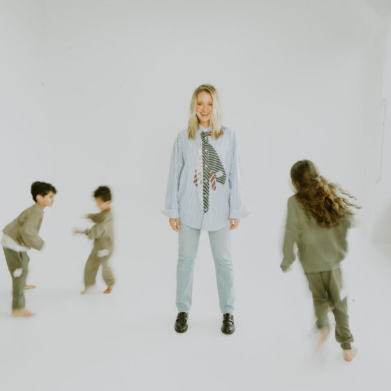 A blonde haired woman standing in the middle of three young children running around her against a white floor and background.