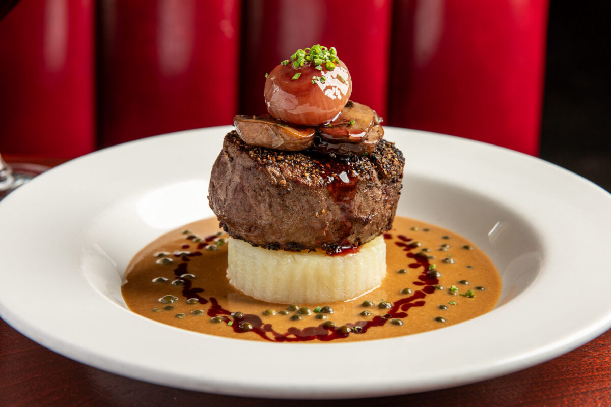 A pepper steak on a white plate with sauce around it and a roasted cherry tomato on top.