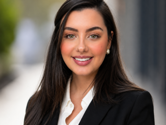 A young woman with long dark hair and well-done makeup is wearing a white top and black blazer. She's looking at the camera and smiling, with her body turned slightly to her right.