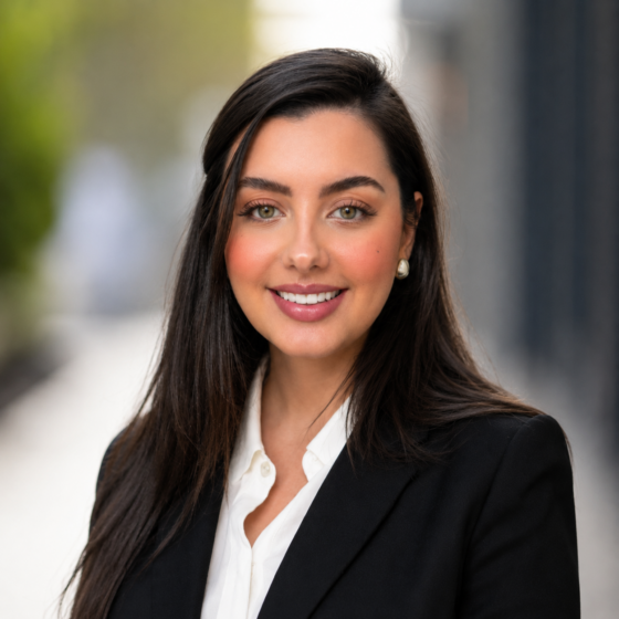 A young woman with long dark hair and well-done makeup is wearing a white top and black blazer. She's looking at the camera and smiling, with her body turned slightly to her right.